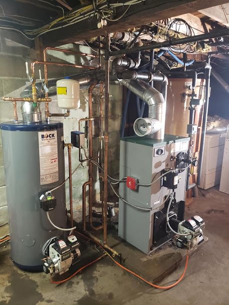 Bock water heater and boiler system installed in a basement with copper piping, circulator pumps, and exhaust ventilation.