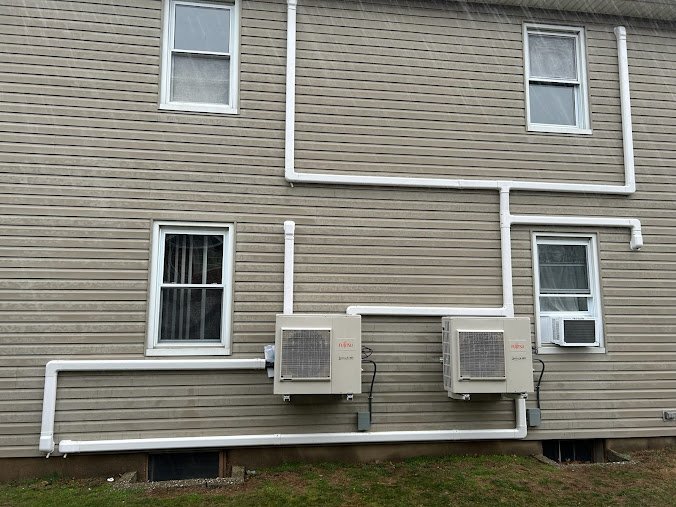 Two Fujitsu mini-split outdoor units mounted on a house with vinyl siding, with extensive line set covers for refrigerant piping.