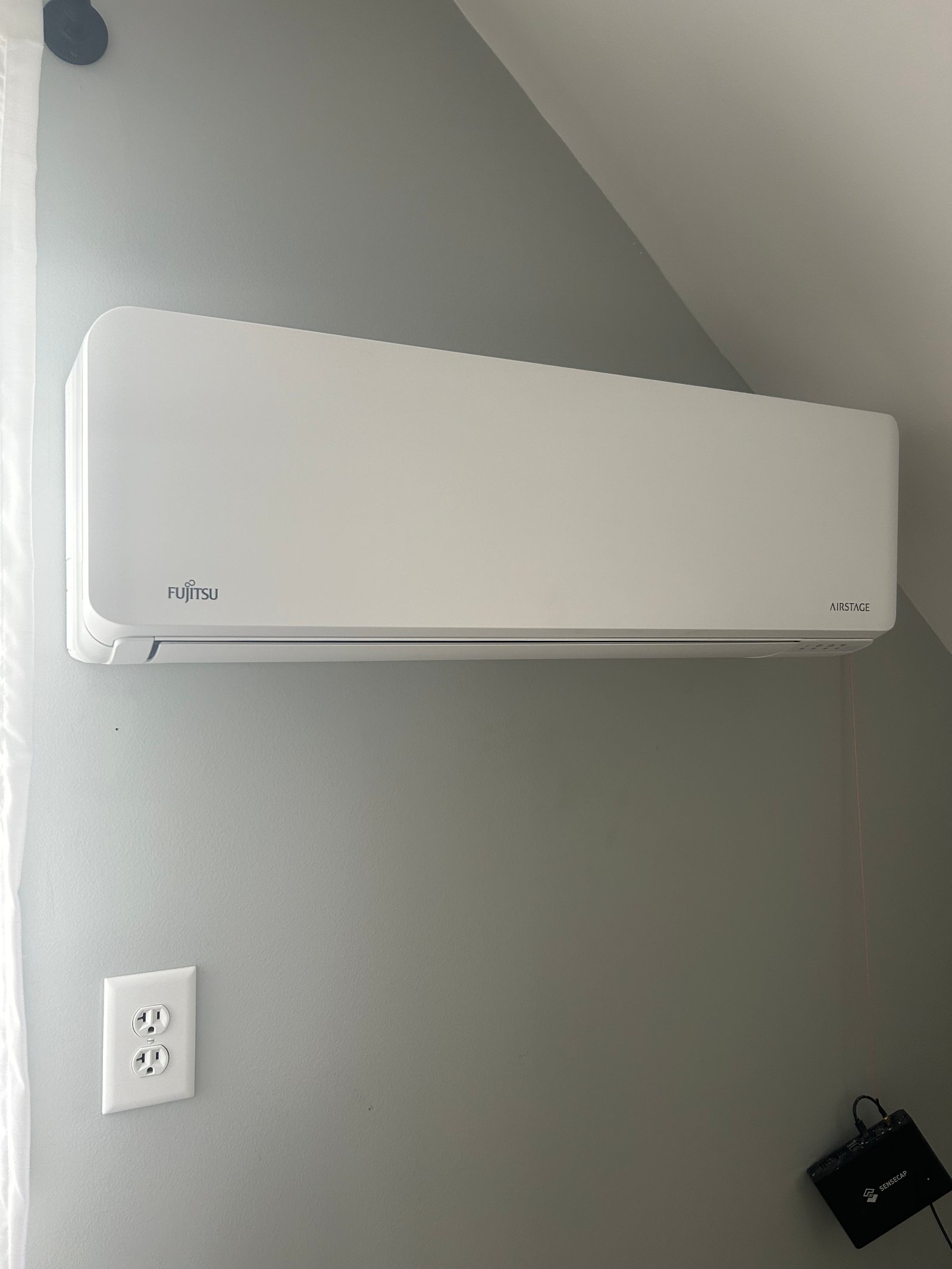 Outdoor installation of a Fujitsu mini-split unit alongside a central air conditioning condenser on the exterior of a house with beige vinyl siding