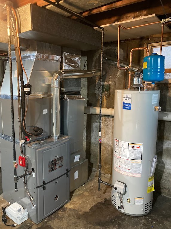 Gas furnace with ductwork and a Bradford White water heater installed in a basement with exposed stone walls and piping.