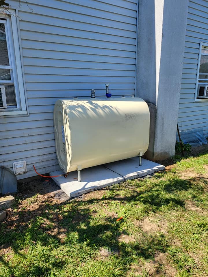 Outdoor heating oil tank installed on concrete pads next to a house with vinyl siding, connected via fuel lines.